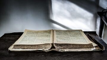 كتاب قديم مفتوح على طاولة خشبية، تظهر الصفحات المصفحة بوضوح. الإضاءة الخافتة تخلق جوًا هادئًا حول الكتاب، مع ظلال تتشكل على الجدران.