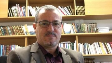 رجل في بدلة رسمية يجلس أمام رفوف مليئة بالكتب في مكتب. يظهر خلفه ترتيب من الكتب والمجلدات، مما يعكس بيئة أكاديمية أو بحثية.