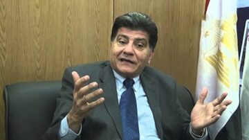 رجل يرتدي بدلة رسمية يجلس على كرسي في مكتب، يتحدث بحماس مع إيماءات يديه، خلفه علم يمثل مصر.