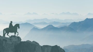 فارس على حصان يقف على حافة صخرية، يطل على مشهد جبلي ضبابي. تتداخل السلاسل الجبلية في الخلفية مع السماء الزرقاء، مما يخلق إحساسًا بالهدوء والاتساع.