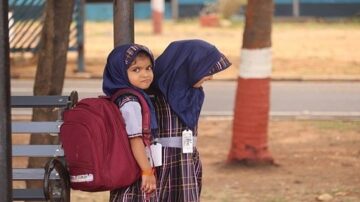 طفلتان يرتديان الزي المدرسي مع حقيبة ظهر حمراء، تقفان بالقرب من شجرة في ساحة المدرسة. تعبيرات وجهيهما تظهر الفضول والبراءة.