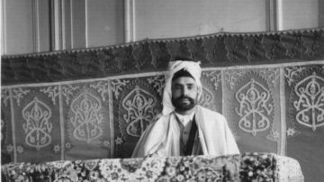 رجل يجلس على وسادة مزخرفة، يرتدي لباس تقليدي مع غطاء رأس، في بيئة مزينة بالزخارف. الخلفية تحتوي على سجادة مزخرفة ونقوش هندسية. تعكس الصورة التراث الثقافي والتقاليد.