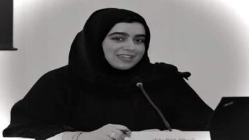 امرأة ترتدي الحجاب تجلس أمام ميكروفون، مبتسمة وتظهر في الخلفية شاشة كمبيوتر. تعبر عن الثقة أثناء مشاركة أفكارها في حدث أو مؤتمر.