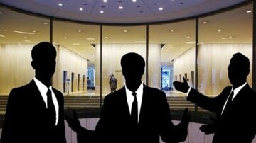 ثلاثة رجال يرتدون بدلات داكنة يقفون في مقدمة مدخل حديث مع جدران زجاجية. يظهر خلفهم شخص واحد داخل المكتب. الإضاءة الساطعة تبرز الفضاء المعاصر.