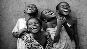 أربعة أطفال يضحكون بفرح، يرتدون ملابس بسيطة، بينما يقفون معًا في وضع غير رسمي. تعكس تعابير وجوههم السعادة والبراءة. الخلفية غير واضحة، مما يبرز تفاعلهم المبهج.