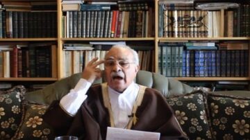 رجل مسن يرتدي ثوبًا تقليديًا يجلس على أريكة مزينة، يتحدث بحماس أمام مكتبة مليئة بالكتب. يرفع إصبعه كإشارة أثناء حديثه، ويبدو أنه يقرأ من ورقة أمامه.