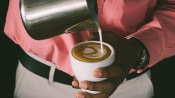 تسريب الحليب في فنجان قهوة، مع تصميم فني على السطح. يظهر شخص يرتدي قميص وردي وسروال فاتح، ممسكًا بكوب أبيض.