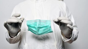 شخص يرتدي قفازات جراحية يحمل كمامة طبية زرقاء. الخلفية بسيطة وغير مزخرفة، مما يبرز أهمية الكمامة في الوقاية الصحية.