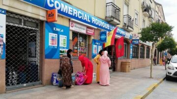 محلات تجارية في حي مزدحم، مع لافتات لعدة خدمات مالية مثل موني جرام. مجموعة من النساء يرتدين ملابس تقليدية، يتجمعن أمام المتجر. تظهر العلم المغربي معروضًا في المدخل، مما يبرز الهوية الثقافية.