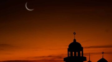 صورة لغروب الشمس مع هلال رقيق يظهر في السماء. silhouetted مآذن مساجد في المقدمة، مع ألوان دافئة من البرتقالي والأحمر في الخلفية.