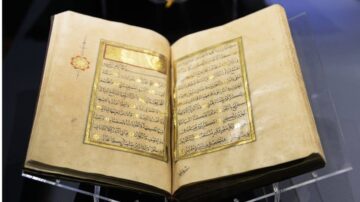 كتاب قديم مفتوح يعرض نصوصًا مكتوبة بخط عربي جميل على صفحات مصنوعة من ورق قديم، مع زخارف ذهبية تضيف لمسة فنية. الكتاب موضوع على حامل زجاجي شفاف، ويظهر تفاصيل فنية دقيقة.