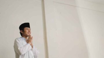 شاب يرتدي ملابس تقليدية إسلامية ويقف مبتسمًا مع يديه مشبكتين في وضعية الصلاة، في غرفة ذات جدران بيضاء وإضاءة طبيعية.