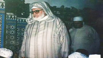 رجل مسن يرتدي جلبابًا تقليديًا مع عمامة، يقف في مكان داخلي مزين بنقوش تقليدية. يظهر خلفه شخص آخر يرتدي ملابس بيضاء. تعكس الصورة أجواء ثقافية ودينية.