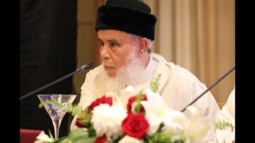 رجل مسن ذو لحية بيضاء يرتدي عمامة سوداء، يتحدث في مؤتمر. أمامه زهور حمراء وبيضاء في فازة، والميكروفون موضوع أمامه.