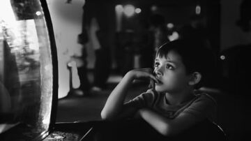 طفل يتأمل بفضول في حوض سمك، مع تعبيرات فضولية على وجهه، في بيئة مظلمة مع خلفية غير واضحة لأشخاص آخرين.