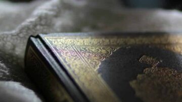 كتاب ذو غلاف مزخرف بتفاصيل ذهبية، موضوع على قماش أبيض، مع خلفية ضبابية تعزز من جمالية الصورة.