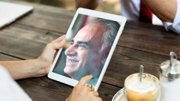 رجل مسن مبتسم يظهر على شاشة جهاز لوحي يُمسك به شخص آخر، مع كوب قهوة على الطاولة. الخلفية تحتوي على طاولة خشبية.