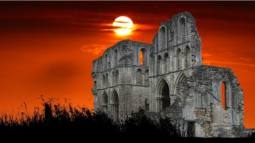 أطلال مبنى قديم أمام غروب الشمس بسماء برتقالية مع أعشاب داكنة في المقدمة.