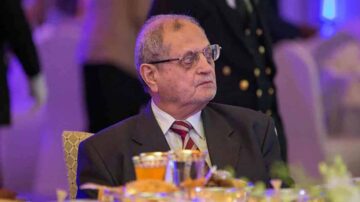 رجل مسن يرتدي بدلة رسمية يجلس على طاولة في مناسبة اجتماعية، مع أطباق طعام ومشروبات أمامه. الإضاءة تتضمن ألوان زاهية، مما يضيف جواً احتفاليًا.