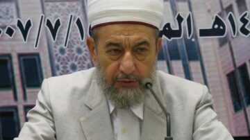 شيخ يرتدي عمامة بيضاء وقبعة حمراء، يجلس أمام ميكروفون ويتحدث بجدية، خلفه لافتة تحمل نصوصًا عربية. يعبّر عن الحكمة والمعرفة في سياق ديني.