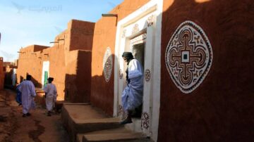 ممر ضيق بين منازل تقليدية مبنية من الطين، يظهر فيه رجل يرتدي ملابس تقليدية، يقف عند باب منزل مزين بنقوش فنية. تتواجد في الخلفية شخصيات أخرى تسير في الشارع، مما يعكس الحياة اليومية في منطقة صحراوية.