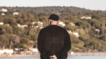 رجل مسن يقف على شاطئ ينظر إلى منظر طبيعي هادئ، مع تلال خضراء ومنازل على خلفية المشهد. الرجل يرتدي قبعة سوداء وسروال داكن، ويداه خلف ظهره.