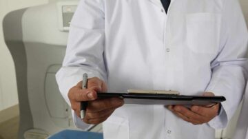 طبيب يرتدي معطفًا أبيض holding clipboard وقلم، في بيئة طبية. يظهر خلفه جهاز طبي، مما يشير إلى إجراء فحص أو استشارة طبية.