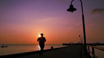 رجل يسير على رصيف بحري أثناء غروب الشمس، مع سماء ملونة بألوان دافئة، وأضواء الشارع مضاءة بشكل خافت، وقوارب في الخلفية.