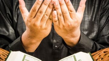 رجل يرتدي قميصًا أسود يحمل القرآن بيد واحدة، بينما يرفع يديه للدعاء. تركز الصورة على تعبير الخشوع والتواصل الروحي.