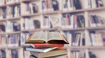 كتب مفتوحة ومكدسة على طاولة أمام رفوف مليئة بالكتب. تشكيلة من الكتب الملونة تظهر في الخلفية، مما يبرز جو المكتبة.