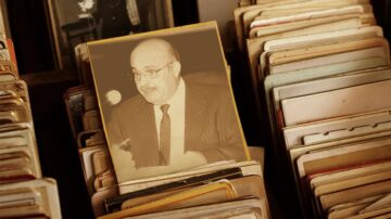 صورة قديمة لرجل يرتدي بدلة، يظهر في إطار مائل بين مجموعة من الصور الفوتوغرافية الموضوع في صندوق. الخلفية مليئة بالصور والبطاقات القديمة، مما يضفي إحساسًا بالحنين إلى الماضي.