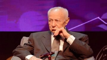 رجل مسن يجلس على أريكة أثناء حدث رسمي، يرتدي بدلة داكنة وقميص أبيض، مع تعبير جاد على وجهه. خلفية الصورة تحتوي على ألوان زاهية وعناصر تصميم حديثة، مما يبرز أهمية المناسبة.