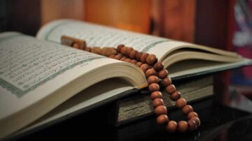 كتاب مفتوح مع سبحة خشبية موضوعة فوق صفحاته، يُظهر زوايا دقيقة للنص العربي. الخلفية ضبابية تضيف جوًا روحانيًا.