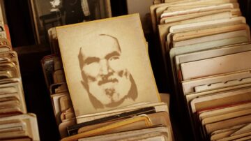 صورة فوتوغرافية قديمة لرجل ذو لحية وشعر أبيض، تظهر على بطاقة وسط مجموعة من البطاقات القديمة المكدسة. تبرز ملامح الوجه بشكل خفيف على خلفية داكنة، مما يضيف طابعاً تاريخياً إلى الصورة.