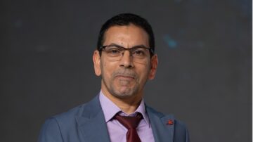 رجل يرتدي بدلة زرقاء وقميص بنفسجي، مع نظارات وقميص بأزرار، يقف أمام خلفية محايدة، مظهره واثق وجاد.