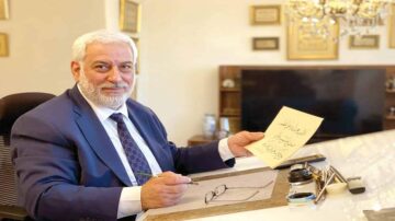 رجل مسن يرتدي بدلة زرقاء يجلس على مكتب أنيق، يحمل ورقة مكتوبة بخط يدوي، ويبتسم. خلفه توجد شاشة تلفاز ومجموعة من الصور والإطارات الزخرفية على الجدران.