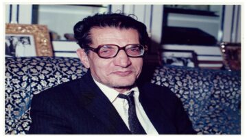 رجل مسن يرتدي نظارات وقميص رسمي مع ربطة عنق، يجلس على أريكة مزخرفة. توجد صور إطارية في الخلفية. تعبير وجهه يظهر الحكمة والتجربة.