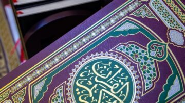 كتاب مزخرف بتصميم تقليدي، يظهر تفاصيل دقيقة باللونين الذهبي والأخضر. الكتاب موضوع على طاولة بجانب مجموعة من الكتب الأخرى. الخلفية تحتوي على سجادة مزخرفة تعكس الطابع الثقافي.