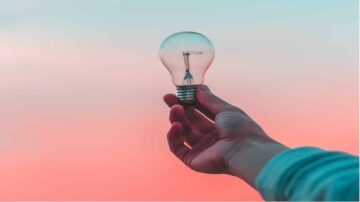 مصباح كهربائي في يد شخص، مع خلفية ملونة تتضمن تدرجات الأزرق والوردي، تعبر عن الإبداع والأفكار الجديدة.