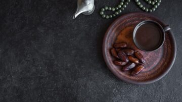 صينية خشبية تحتوي على تمور بجانب فنجان قهوة على سطح داكن، مع إبريق فضي وزخرفة من الخرز.