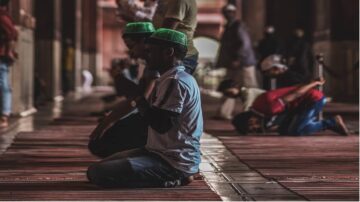 شخص يجثو في مسجد أثناء الصلاة، يرتدي قبعة خضراء. خلفه مجموعة من المصلين في أوضاع مختلفة للصلاة، مع أجواء هادئة وإضاءة خافتة. يعكس المشهد الروحانية والتقاليد الإسلامية.