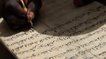 يد تظهر كتابة نص بخط يدوي على ورقة قديمة. الحبر الأسود يبرز على الخلفية الفاتحة للورقة. الكتابة تبدو تقليدية، مما يعكس التراث الثقافي.