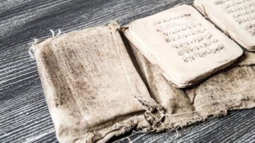 كتاب قديم مع صفحات متسخة وممزقة، موضوعة على سطح خشبي داكن. يظهر الكتاب علامات الاستخدام والتآكل، مما يدل على قدمه وأهميته الثقافية.