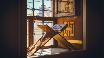 مصحف موضوع على حامل خشبي مزخرف بجانب نافذة، مع إضاءة طبيعية تتسلل من خلالها، تعكس تفاصيل هندسية جميلة في الخلفية.