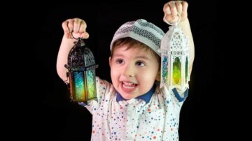 طفل مبتسم يحمل فانوسين ملونين في يديه. يرتدي قبعة بيضاء وقميص مزخرف بنقاط ملونة. الخلفية داكنة، مما يبرز الفوانيس.