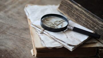 عدسة مكبرة موضوعة على رسائل قديمة مربوطة بخيوط، مع كتاب قديم في الخلفية. تعكس الصورة جوًا من الاستكشاف والبحث عن المعرفة التاريخية.