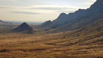 منظر طبيعي لصحراء واسعة مع تلال وجبال في الخلفية، السماء صافية مع بعض السحب. الأرض جافة ومغطاة بالأعشاب القصيرة، مما يبرز جمال الطبيعة القاحلة.