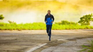 رجل يجري على طريق مع خلفية طبيعية خضراء وضباب خفيف، يرتدي سترة زرقاء وسروال رياضي. يعكس النشاط والحيوية في بيئة هادئة.