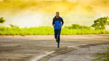 رجل يجري على طريق مع خلفية طبيعية خضراء وضباب خفيف، يرتدي سترة زرقاء وسروال رياضي. يعكس النشاط والحيوية في بيئة هادئة.