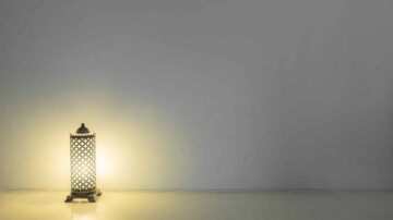 مصباح مزخرف ينبعث منه ضوء دافئ، موضوع على سطح أملس بجانب جدار فارغ. يخلق الإضاءة المحيطة جوًا مريحًا.